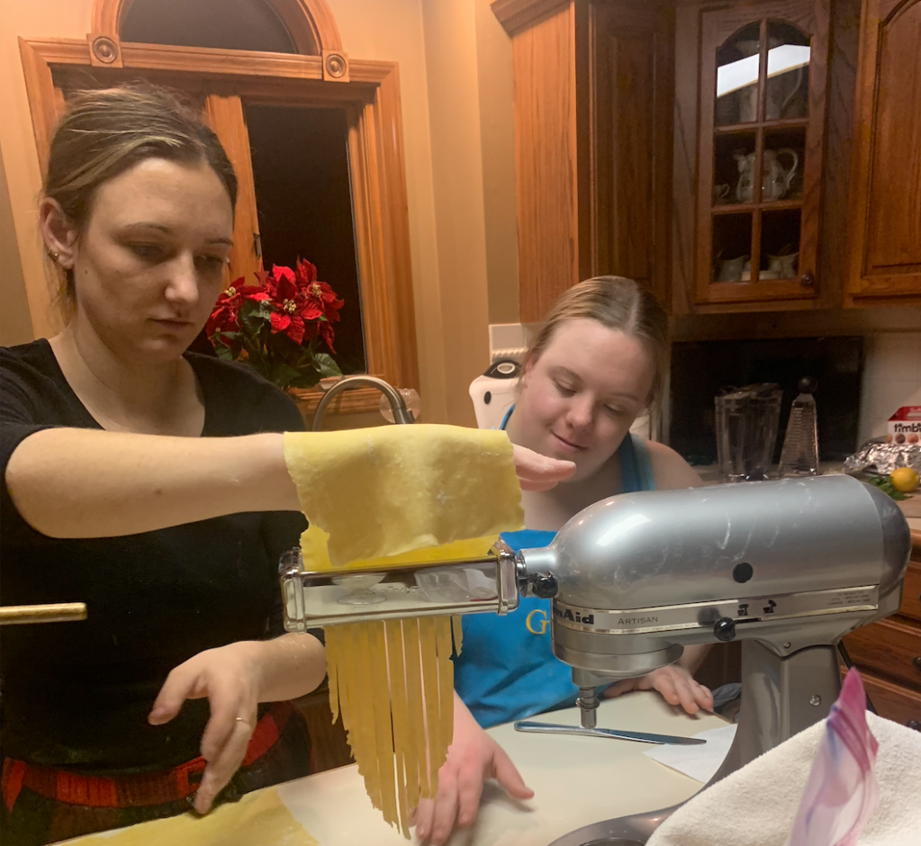 image description: Lauren puts the flat pasta dough through the fettuccine maker and Genna catches the pieces on the bottom.