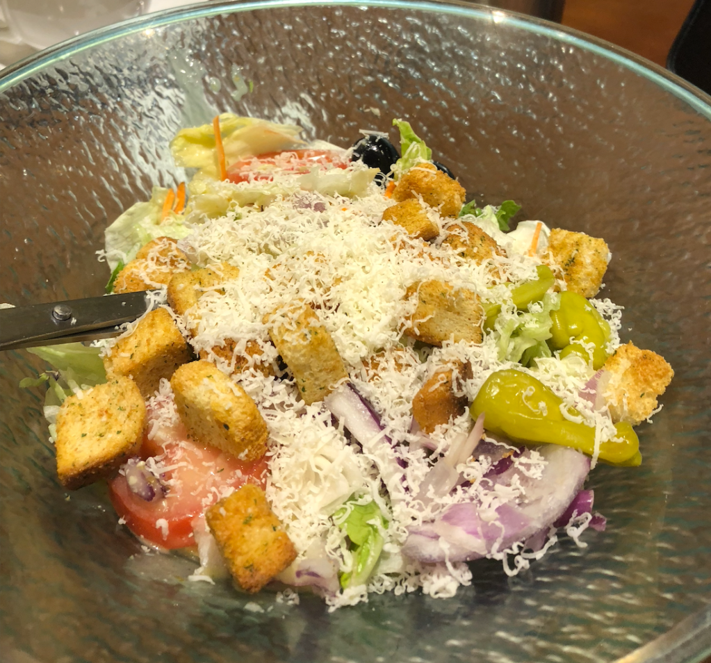 image description: a bowl of salad that has croutons, parmesan cheese, lettuce, peppers, tomatoes, lettuce, and onion from The Olive Garden.