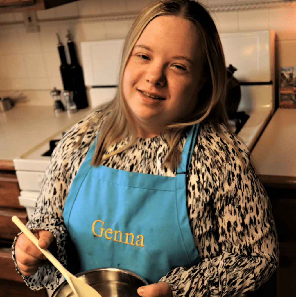 image description: Chef Genevieve stands in her kitchen wearing a blue apron that says 'Genna' and holding a spoon and bowl.